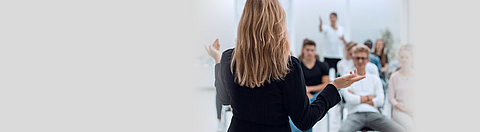 Eine Frau von hinten, die einer sitzenden Gruppe im Hintergrund etwas erklärt.