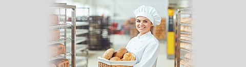 Bäckerin steht lächelnd mit Brotkorb in der Hand in der Backstube
