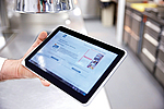 Hand hält Tablet mit OrgaCheck auf dem Bildschirm, im Hintergrund eine Küche
