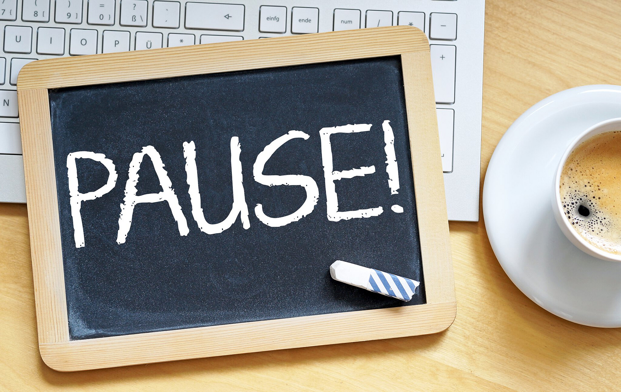 Auf dem Tisch steht eine Tasse Kaffee, eine Tastatur und eine Tafel. Auf der Tafel steht Pause