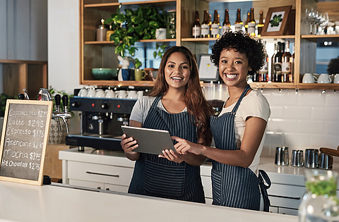 Zwei Frauen arbeiten im Bistro hinterm Tresen und schauen auf ein Tablet.