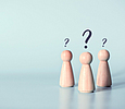 Über drei Holzspielfiguren schweben Fragezeichen, hellblauer Hintergrund