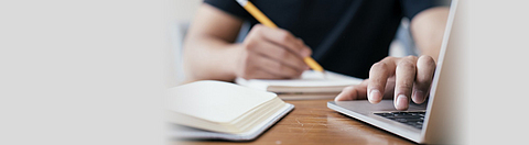 Jemand schreibt mit einem Stift auf einem Schreibblock und tippt mit der anderen Hand auf eine Laptop-Tastatur.