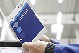 Mann hält Vorschrift in der Hand, im Hintergrund Produktionshalle