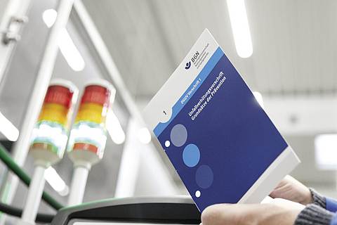 Mann hält Vorschrift in der Hand, im Hintergrund Produktionshalle