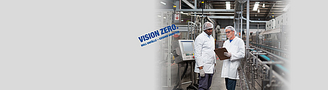 Zwei Männer stehen in einer Fabrikhalle und schauen auf ein Klemmbrett.