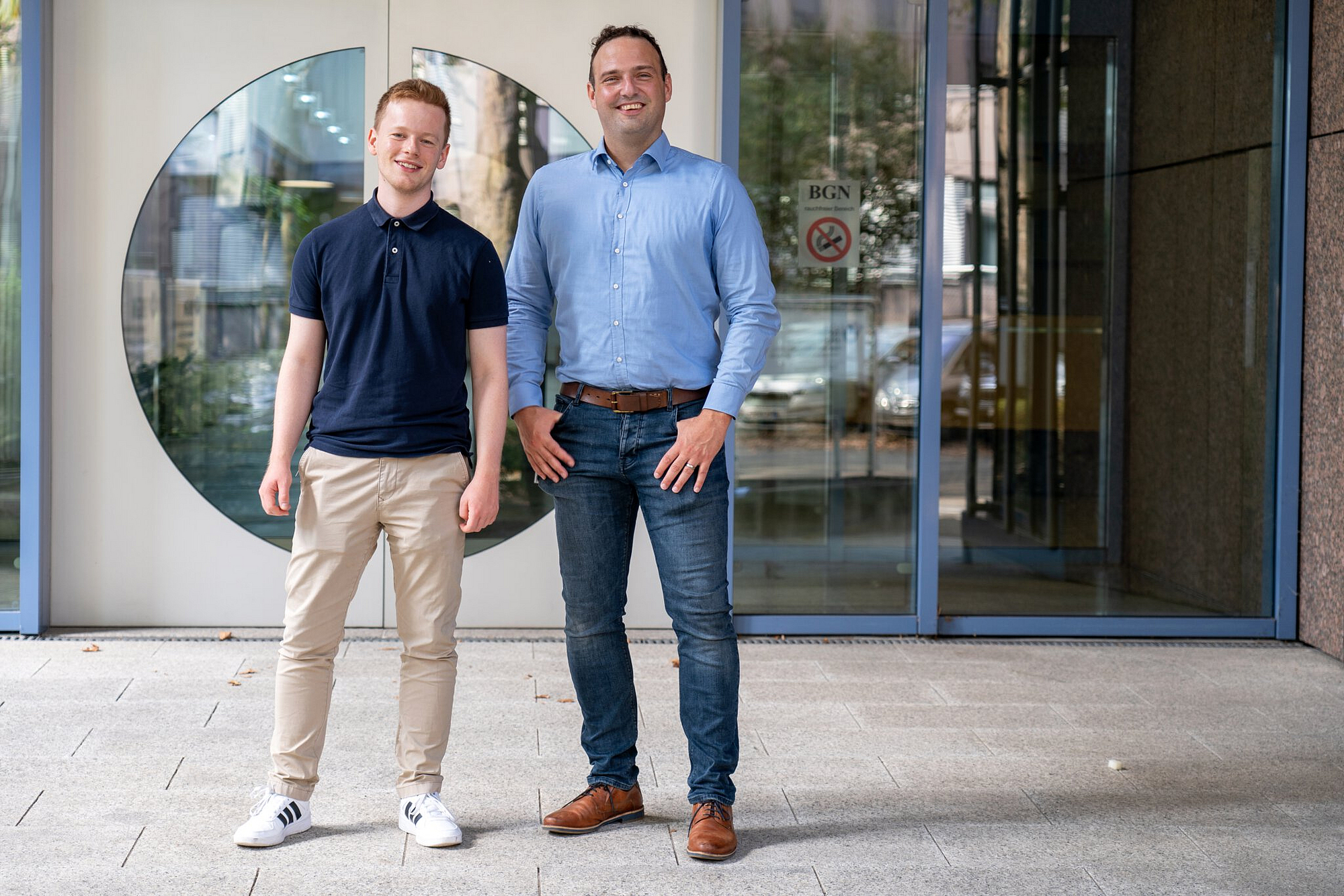 Von links: Jonas Brenneis und Dr. Andre Becker stehen lächelnd vor der Eingangstür des BGN-Schulungszentrums