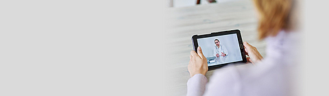 Frau schaut auf ein Tablet, das einen Arzt per Videochat zeigt.