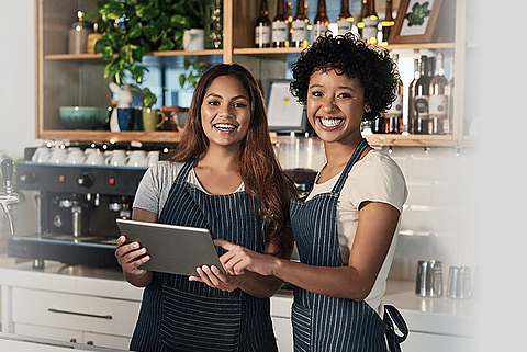 Zwei lächelnde Frauen stehen im Bistro hinterm Tresen und schauen auf ein Tablet.
