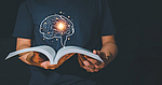 Eine Person liest ein Buch. Aus dem Buch schwebt ein gezeichnetes Gehirn, das in der Mitte hell aufleuchtet.