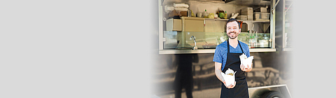 Ein Verkäufer steht vor einem Foodtruck und hält Essen in die Kamera.