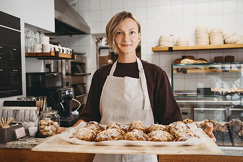 Bäckerin steht im Laden und hält ein Blech voller Croissants in der Hand.