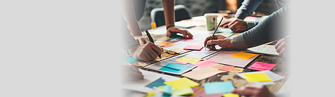 Verschiedene Menschen stehen um einen Tisch voller Dokumente und bunter Notizzettel und schreiben Ideen auf.