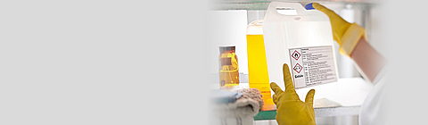 Jemand hebt mit gelben Gummihandschuhen einen Kanister mit Gefahrensymbolen hoch.
