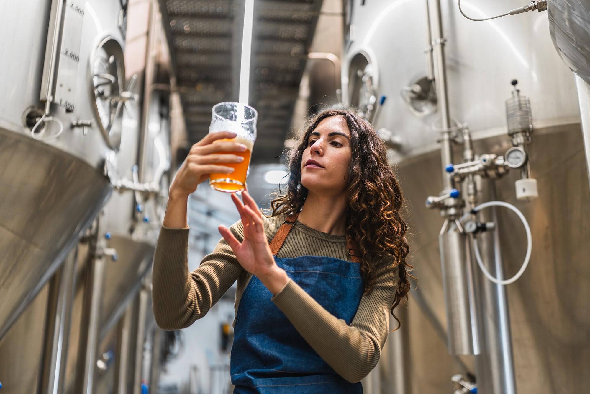 Braumeisterin hält ein Glas Bier und prüft die Qualität zwischen großen Edelstahlbehältern.
