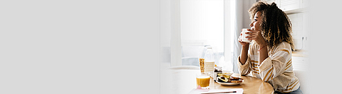 Frau isst an einem Tisch und schaut nachdenklich aus dem Fenster.