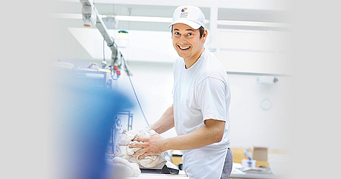 Ein Bäcker knetet Teig und lacht in die Kamera.